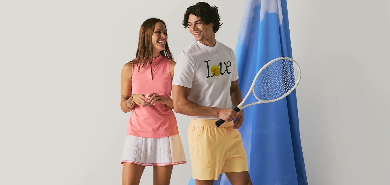 Un uomo e una donna sorridenti in abbigliamento da tennis stanno vicino a uno sfondo di tessuto blu, con l'uomo che tiene una racchetta da tennis.