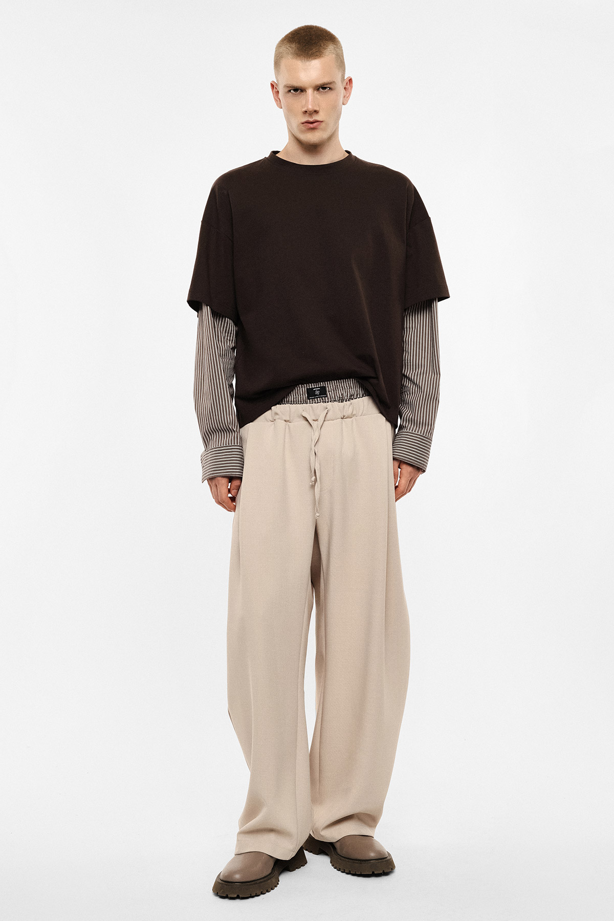 Male model wearing oversized black T-shirt layered over striped long-sleeve shirt, wide beige trousers and brown shoes