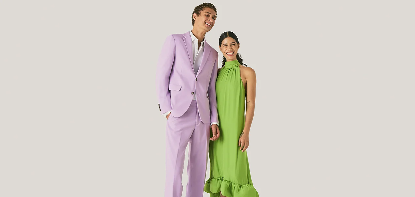 Un uomo in un abito viola chiaro e una donna in un abito senza maniche verde brillante stanno sorridendo contro uno sfondo chiaro e semplice.