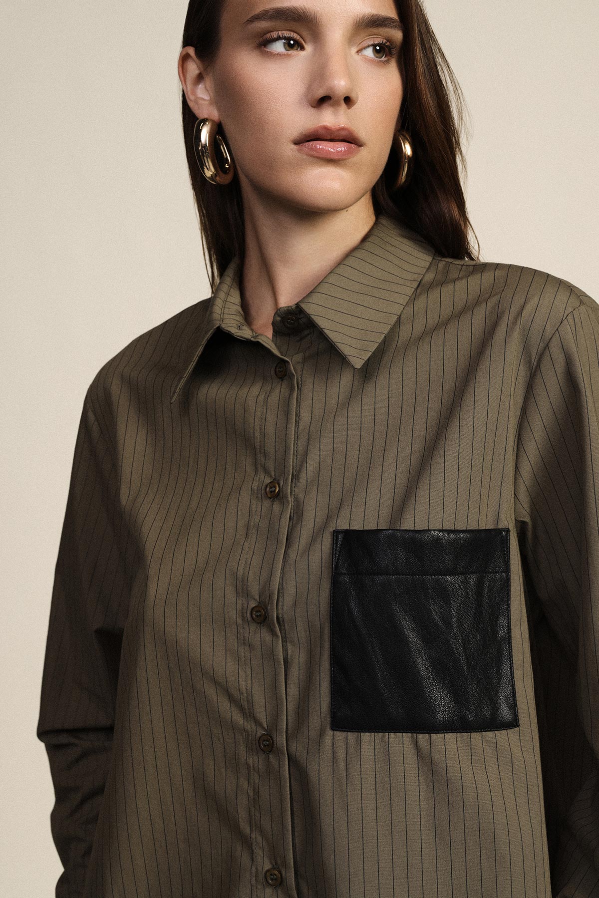 Woman wearing khaki pinstripe button-up shirt with black leather chest pocket and large gold hoop earrings, looking right