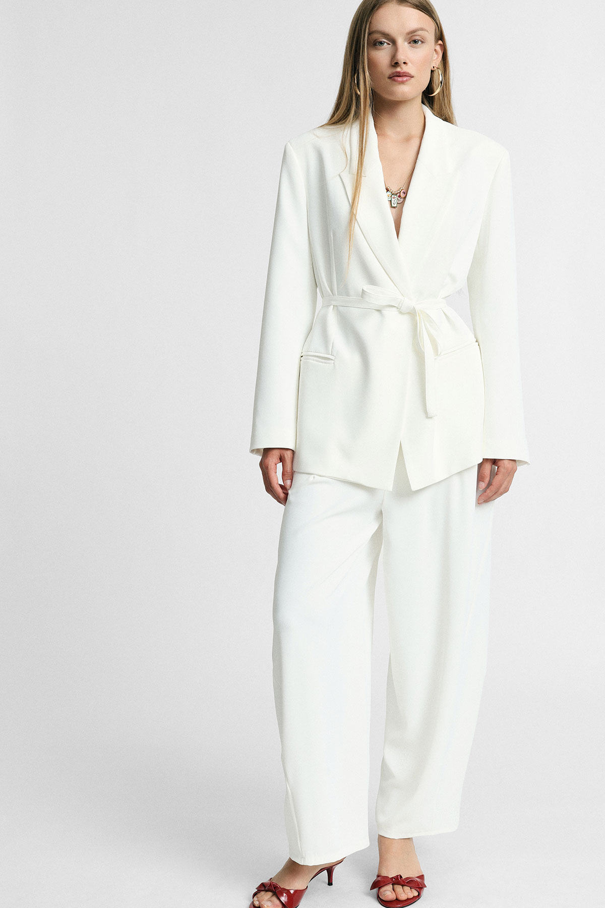 Woman modelling white belted tailored blazer and wide-leg trousers with red heeled sandals on a neutral background