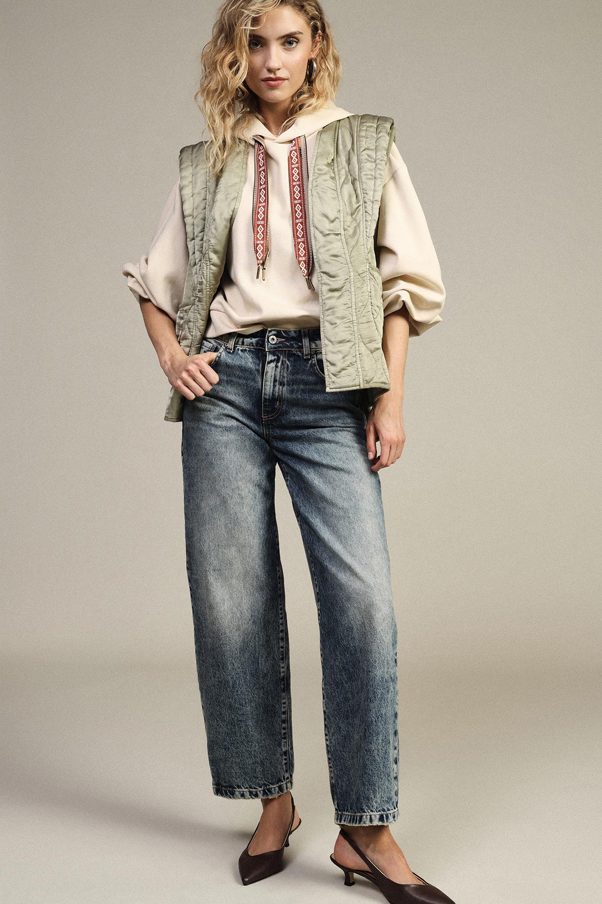 Woman in beige hoodie and green quilted gilet, wide-leg faded jeans and brown slingback heels, standing with hand on hip