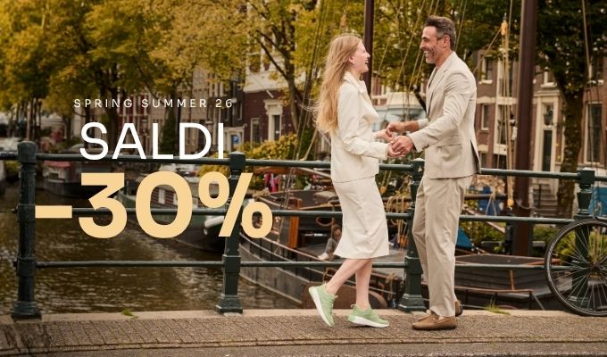 Couple in light spring outfits holding hands and laughing on a canal bridge beside boats with 'Saldi -30%' sale graphic