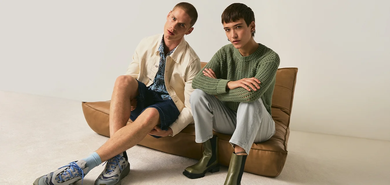 Due giovani adulti siedono su un divano a pavimento in pelle marrone; uno indossa una giacca color crema e pantaloncini, l'altro un maglione verde con pantaloni chiari e stivali.