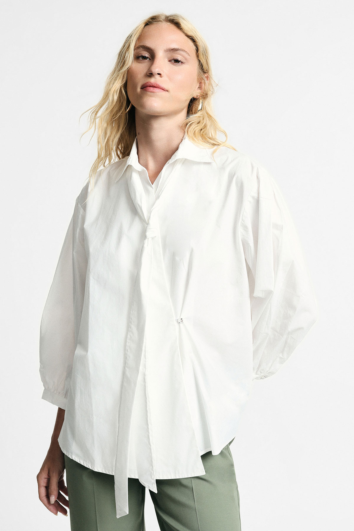 Model wearing oversized white tie-front blouse with balloon sleeves and sage-green trousers against plain white background