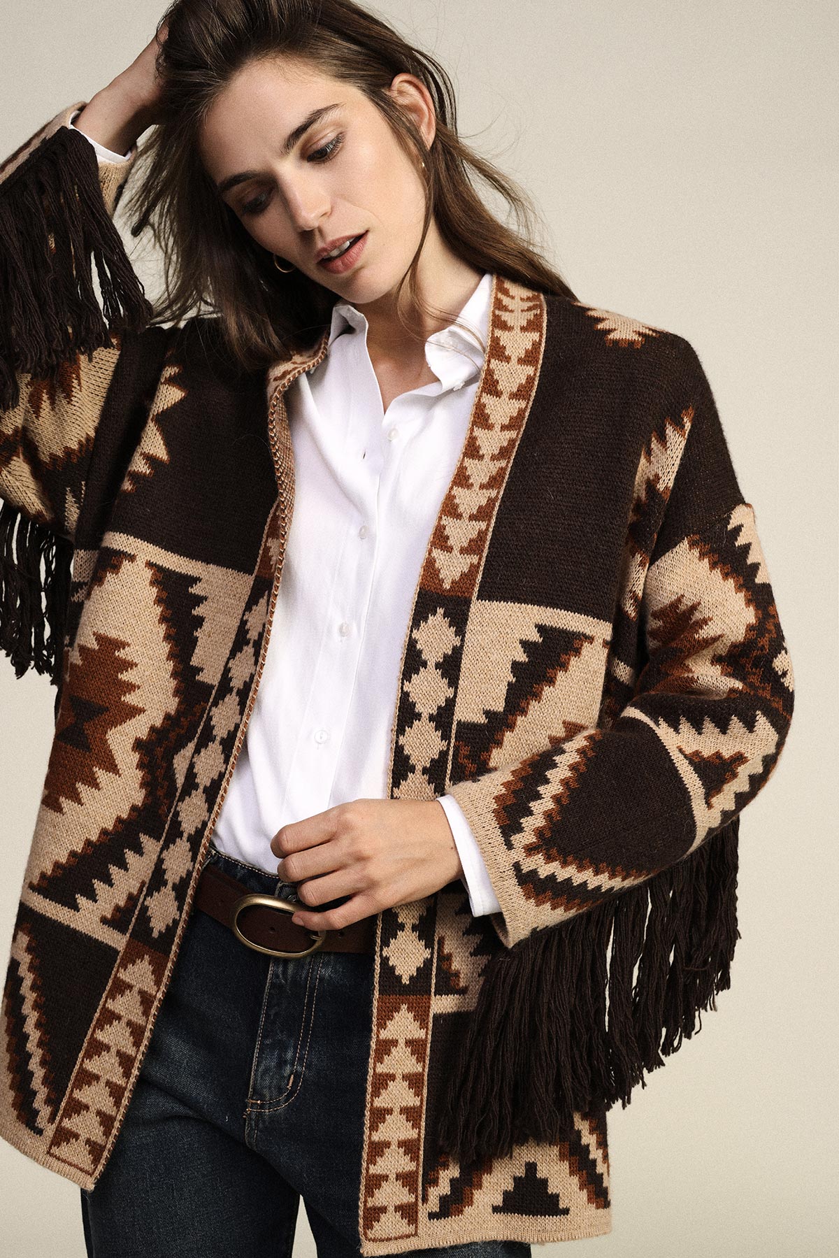 Woman wearing brown and beige Aztec-patterned fringed cardigan over white shirt and dark jeans, adjusting belt