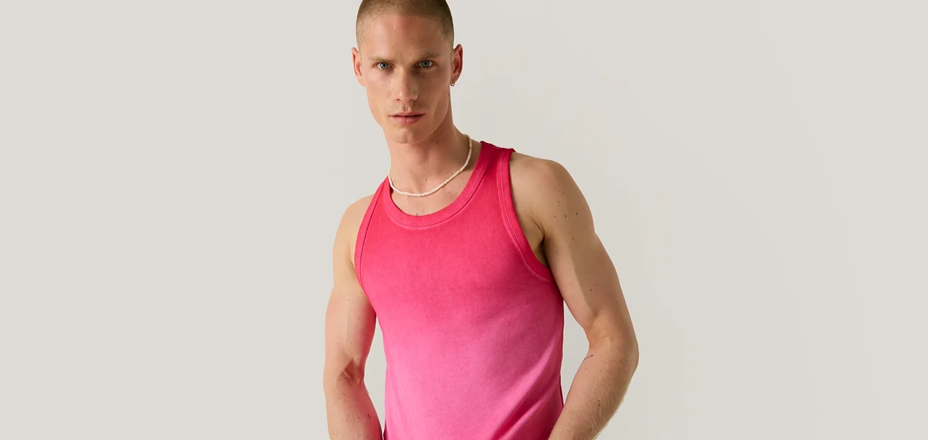 Uomo con capelli corti che indossa una canottiera rosa sfumata senza maniche e pantaloni beige, in piedi davanti a uno sfondo grigio chiaro uniforme.