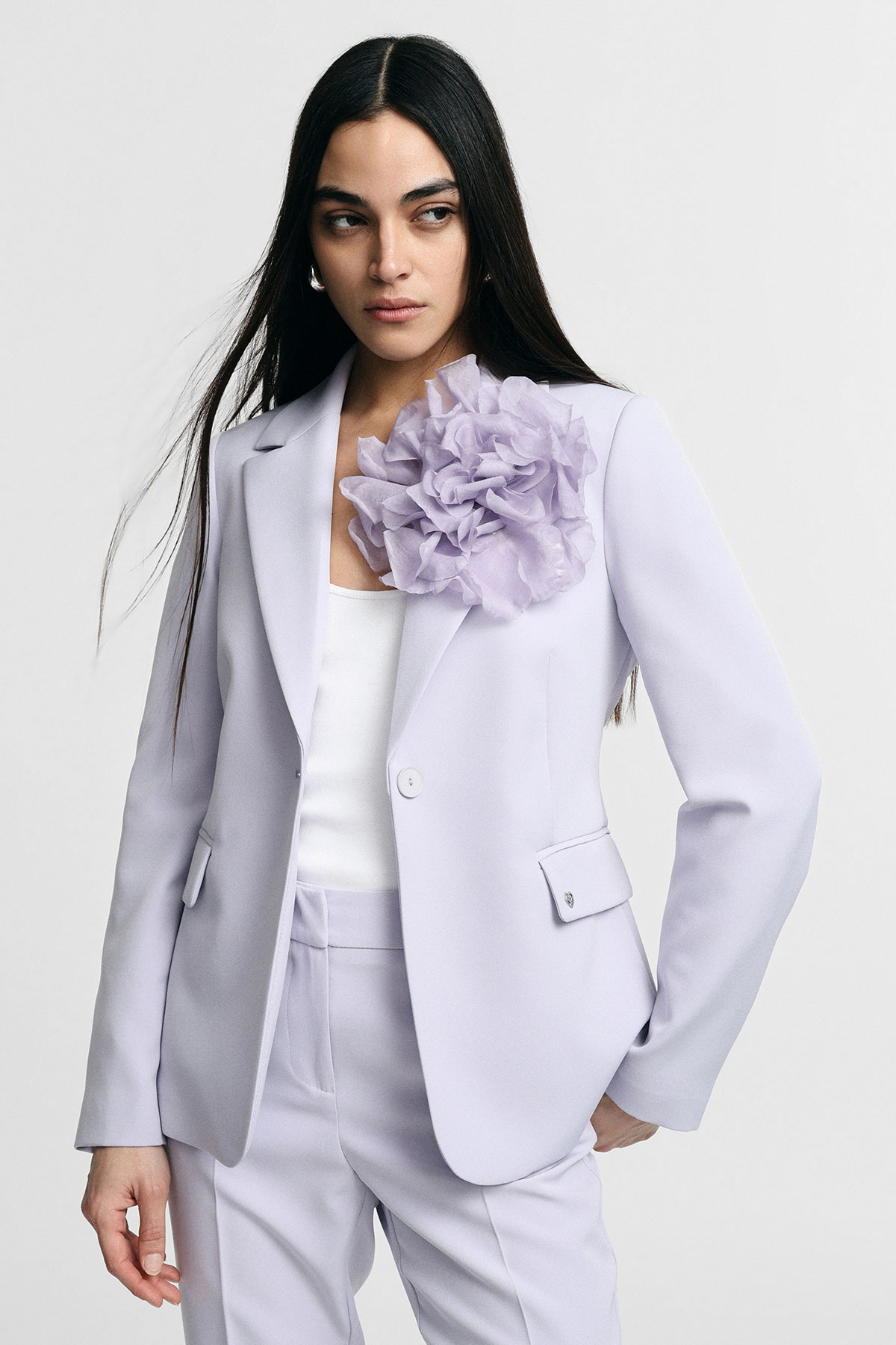 Woman wearing a pale lilac suit with an oversized fabric flower on the lapel and a white top, long dark hair, looking to the side
