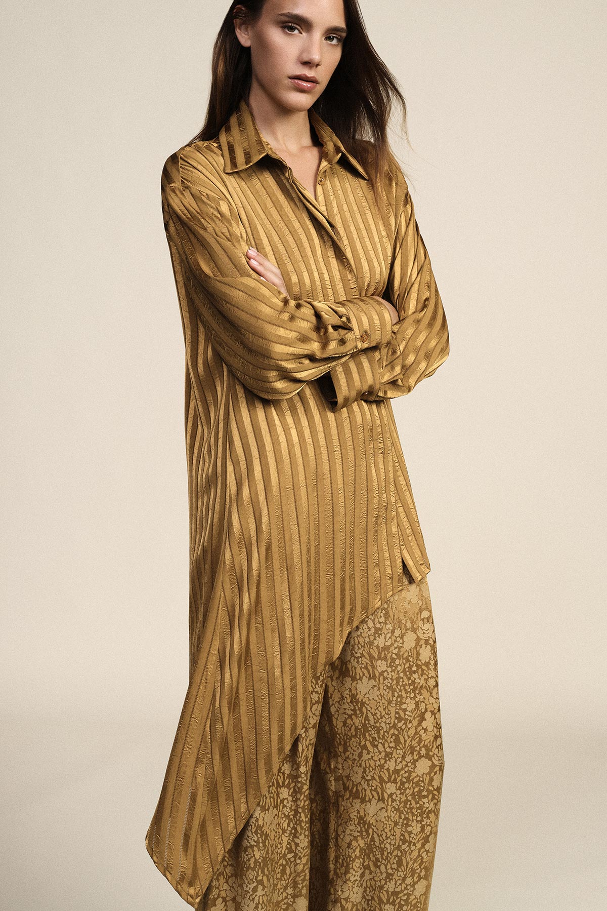 Woman wearing gold satin striped long shirt dress over patterned wide-leg trousers, arms crossed against a neutral background