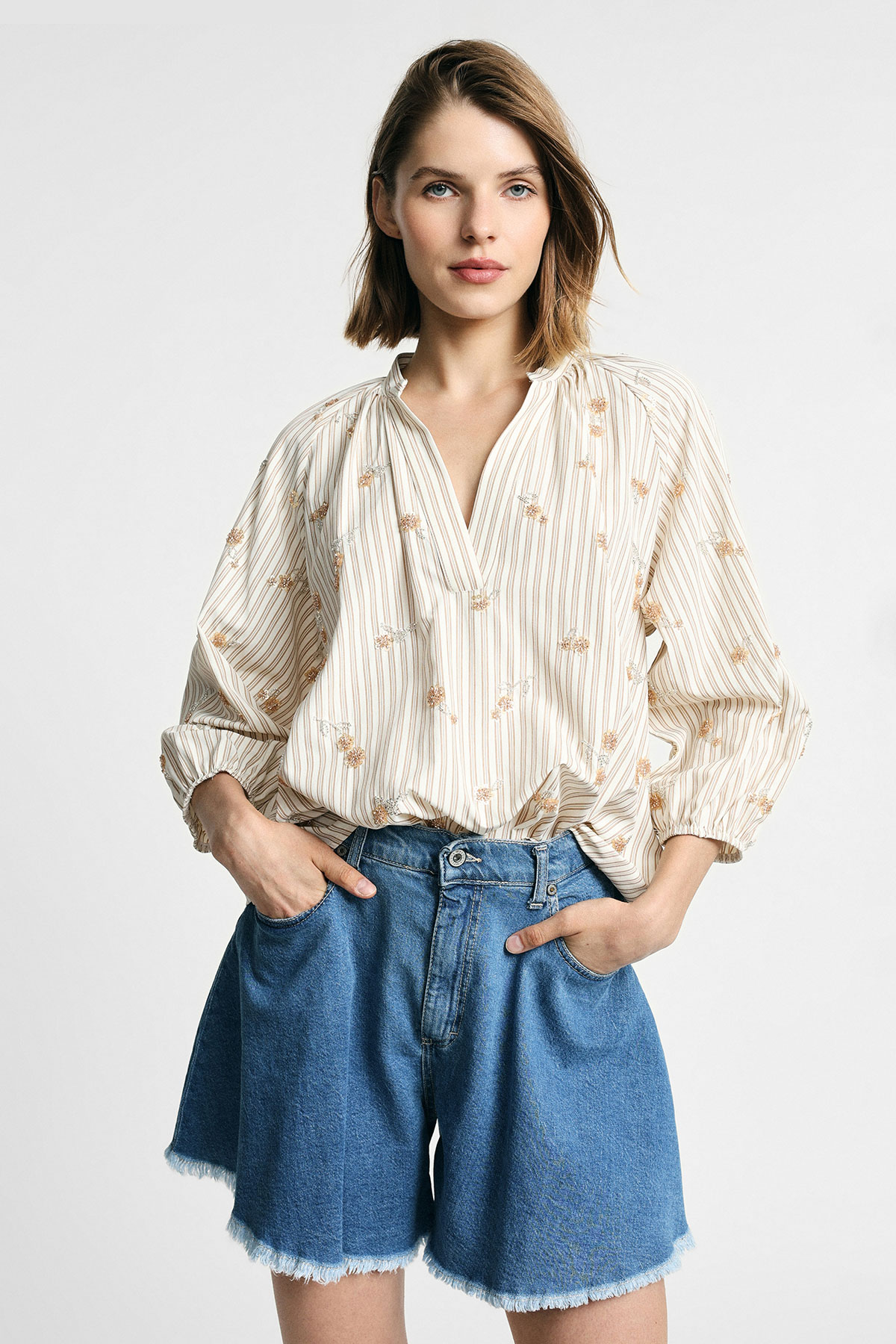 Woman wearing cream striped embroidered blouse tucked into high-waist frayed-hem denim shorts, hands in pockets