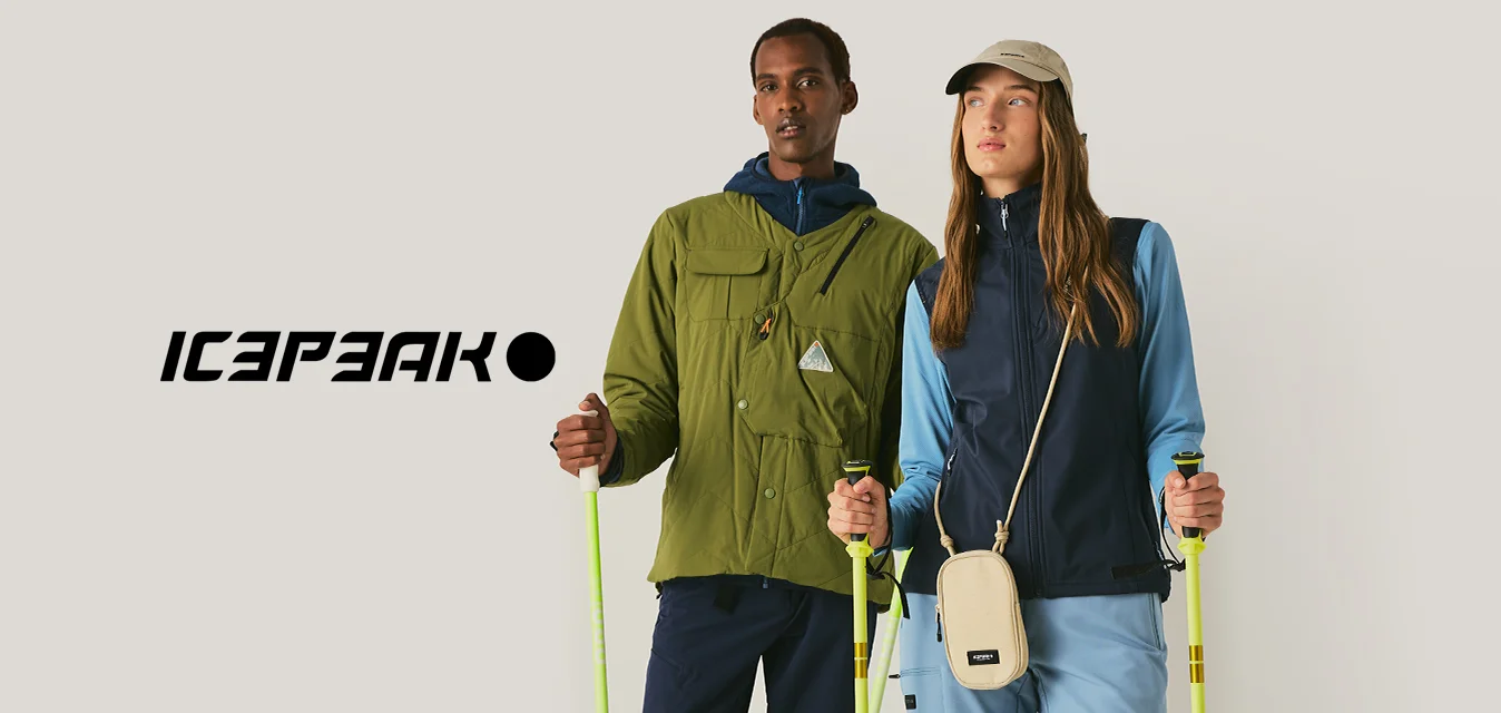 Due persone con abbigliamento da esterno e che tengono bastoncini da trekking sono in piedi davanti a uno sfondo semplice, rivolte in avanti.