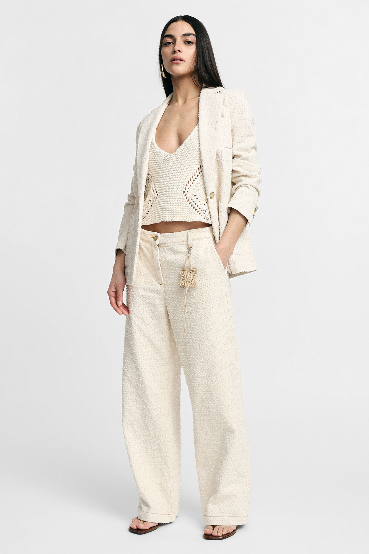 Woman modelling cream textured blazer, knitted crop top and wide-leg trousers with tassel charm, standing on white background