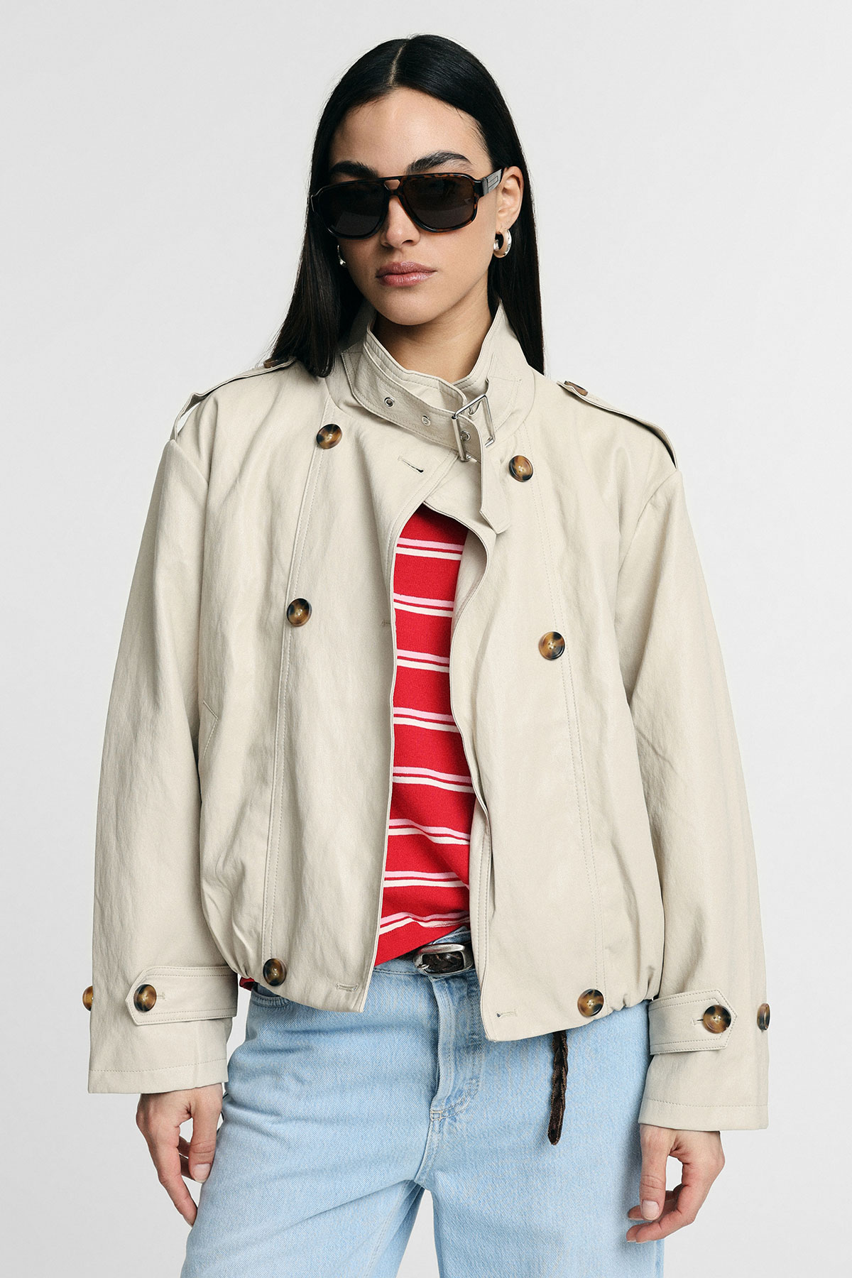 Woman wearing beige cropped trench jacket, red striped top, light blue jeans and dark sunglasses on plain grey background