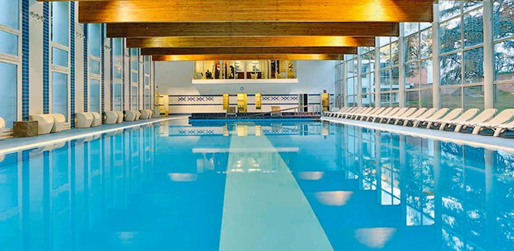 Indoor hotel swimming pool with clear blue water, centre lane, wooden ceiling beams and rows of white loungers beside floor-to-ceiling windows