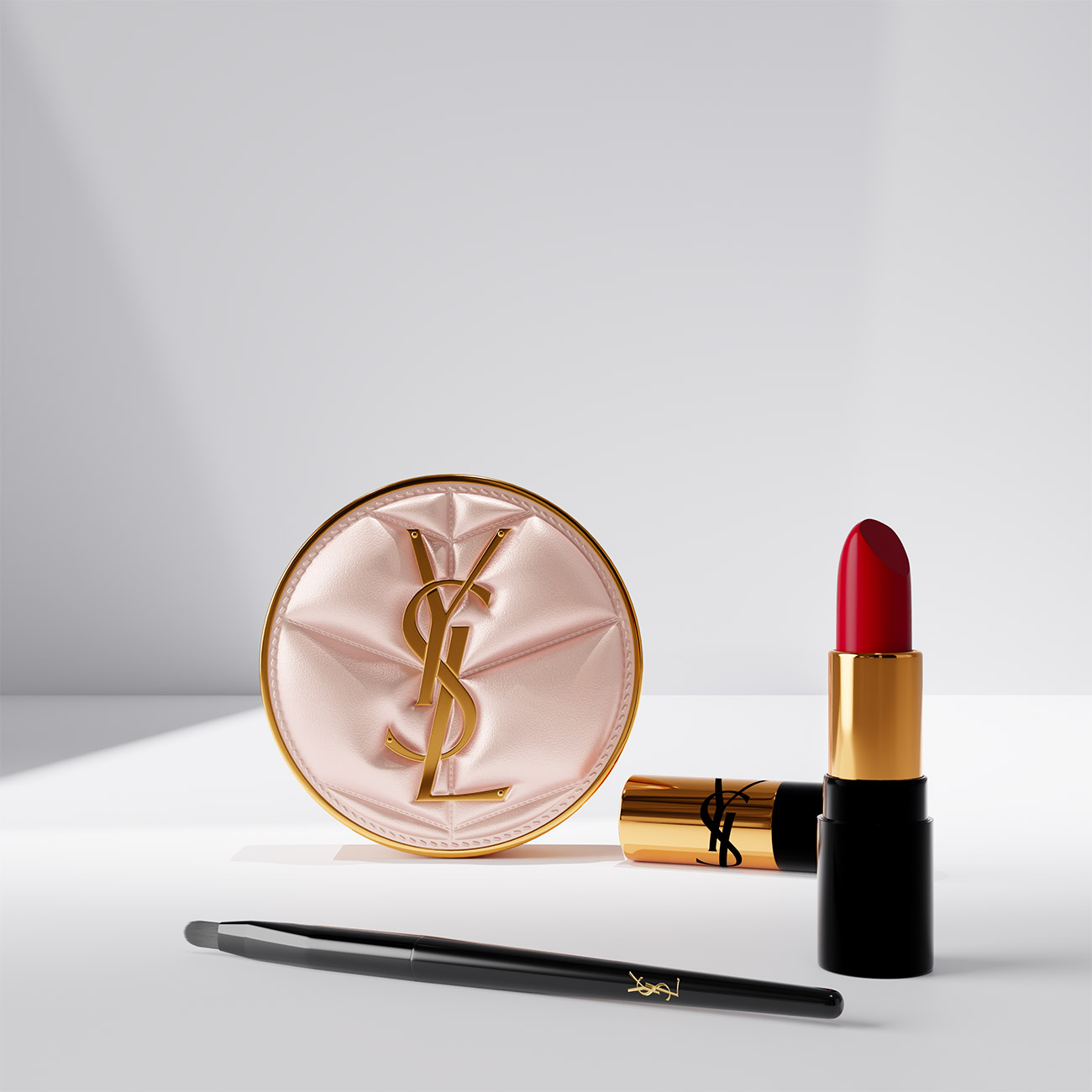 Un compatto cushion rosa trapuntato YSL, un rossetto rosso e un pennello nero esposti su una superficie bianca.