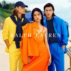 Tre modelli stanno su una spiaggia. Uno indossa un completo giallo con un maglione blu scuro, il secondo un vestito arancione con una giacca coordinata, e il terzo un completo blu.