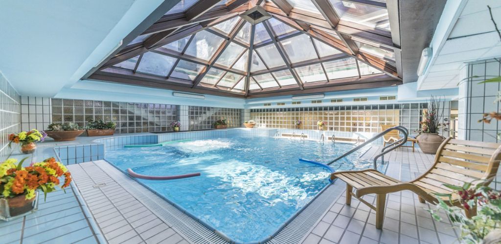 Indoor hotel swimming pool with glass atrium roof, bubbling water jets, wooden deck chairs and potted plants
