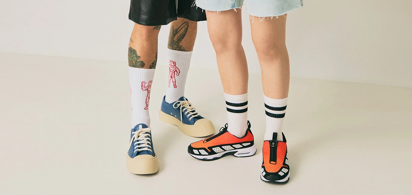 Due persone in piedi su un pavimento chiaro, una indossa pantaloni corti neri, scarpe da ginnastica blu e calzini bianchi con disegni rossi, l'altra indossa pantaloni corti in denim, scarpe da ginnastica arancioni e calzini bianchi a righe.