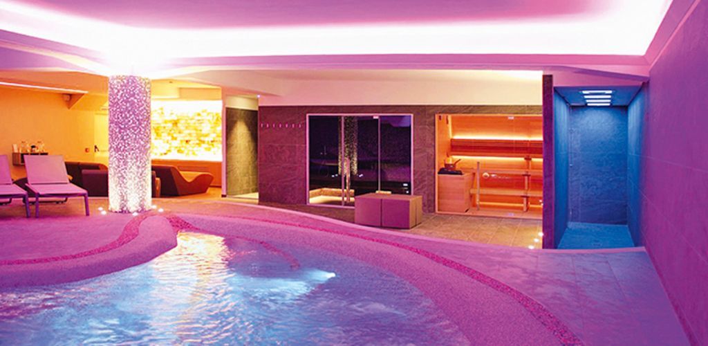 Luxury indoor spa pool with purple-pink mood lighting, lounge chairs and sauna room.
