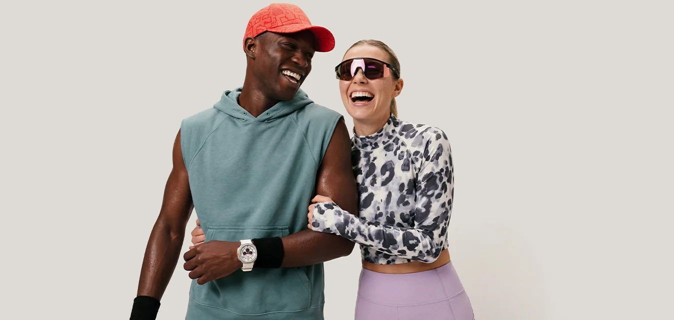 Uomo sorridente con cappellino rosso e felpa senza maniche che si intreccia con una donna che ride con top fantasia e leggings viola su sfondo neutro.