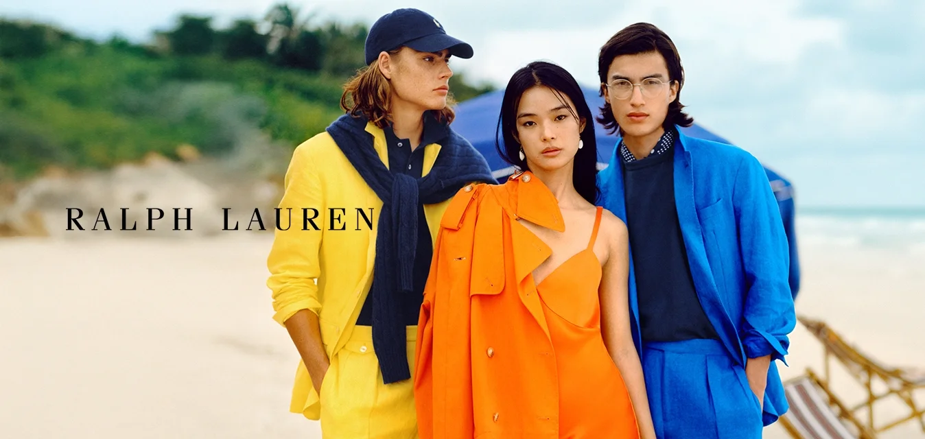 Tre modelli stanno su una spiaggia. Uno indossa un completo giallo con un maglione blu scuro, il secondo un vestito arancione con una giacca coordinata, e il terzo un completo blu.