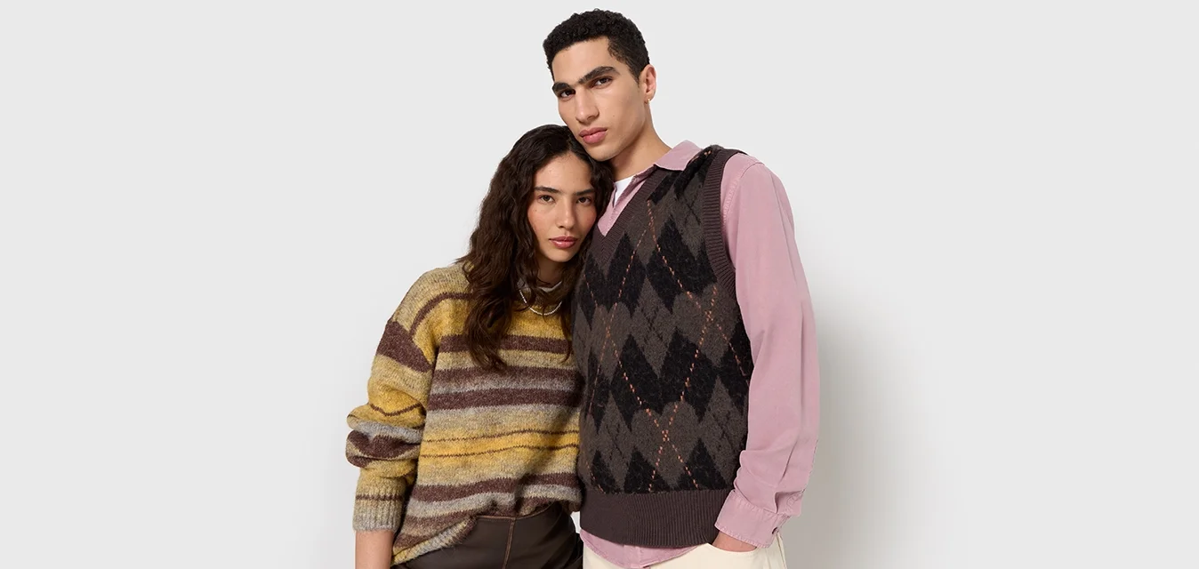 Maglione a righe in giallo, marrone e grigio. Indossato sopra una camicia rosa con colletto. Gilet lavorato a maglia con motivo a diamante in grigio scuro.