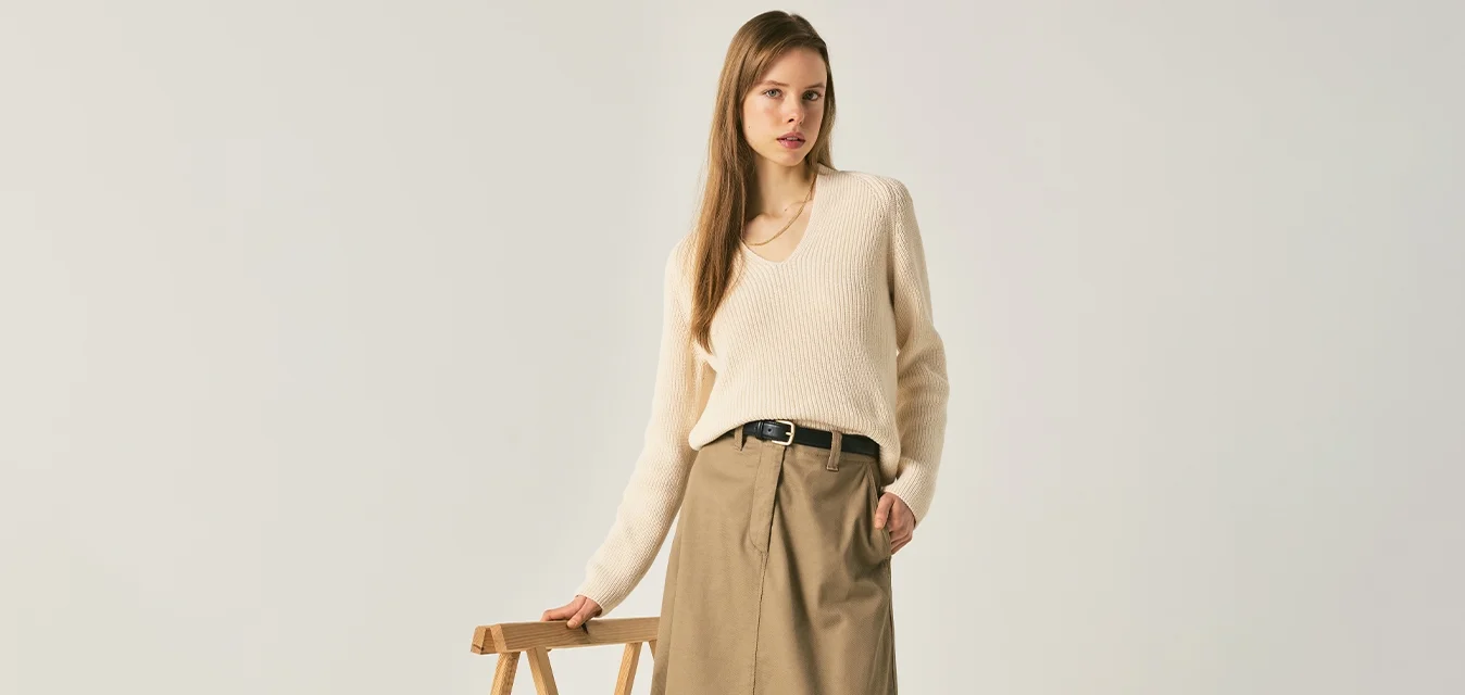 Donna con maglione panna, gonna midi beige e mocassini neri, in piedi all'interno mentre tiene un cavalletto di legno con una mano.