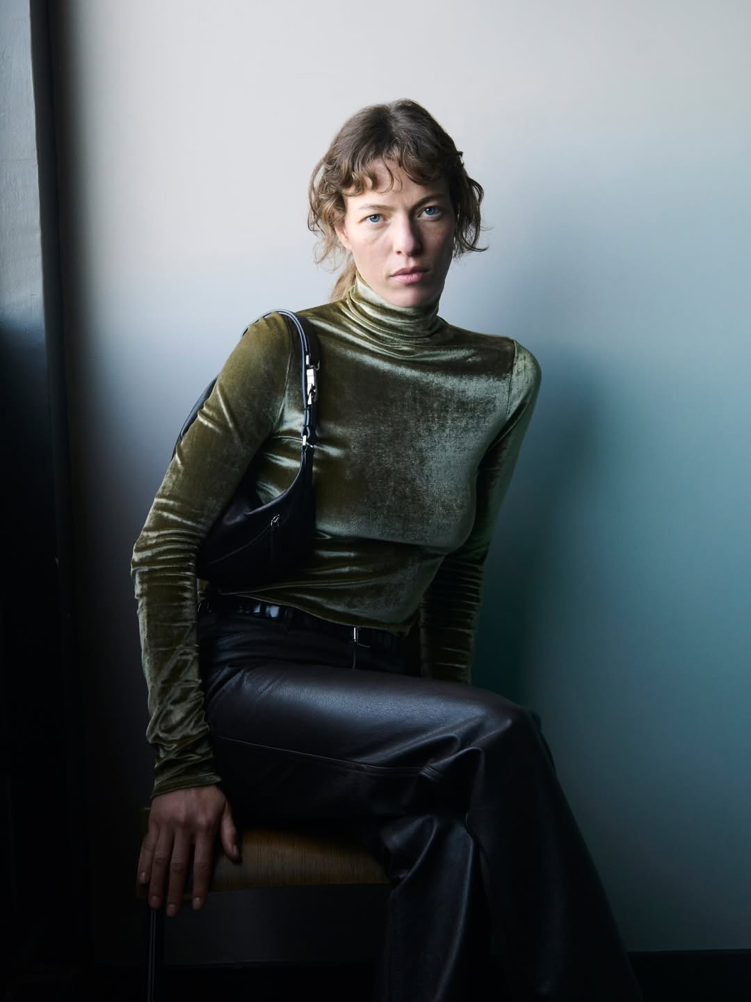 Seated person in olive velvet turtleneck and black leather trousers with a small shoulder bag against a neutral background