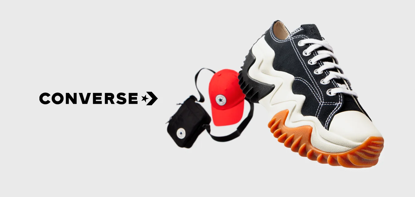 Sneaker bianco e nero con suola a zigzag e dettagli arancioni, accanto a un cappellino rosso e a una piccola borsa nera con un logo circolare.