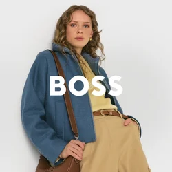 Donna che indossa una giacca blu, pantaloni beige e un top giallo, mentre tiene una borsa a tracolla marrone, in piedi davanti a uno sfondo semplice.