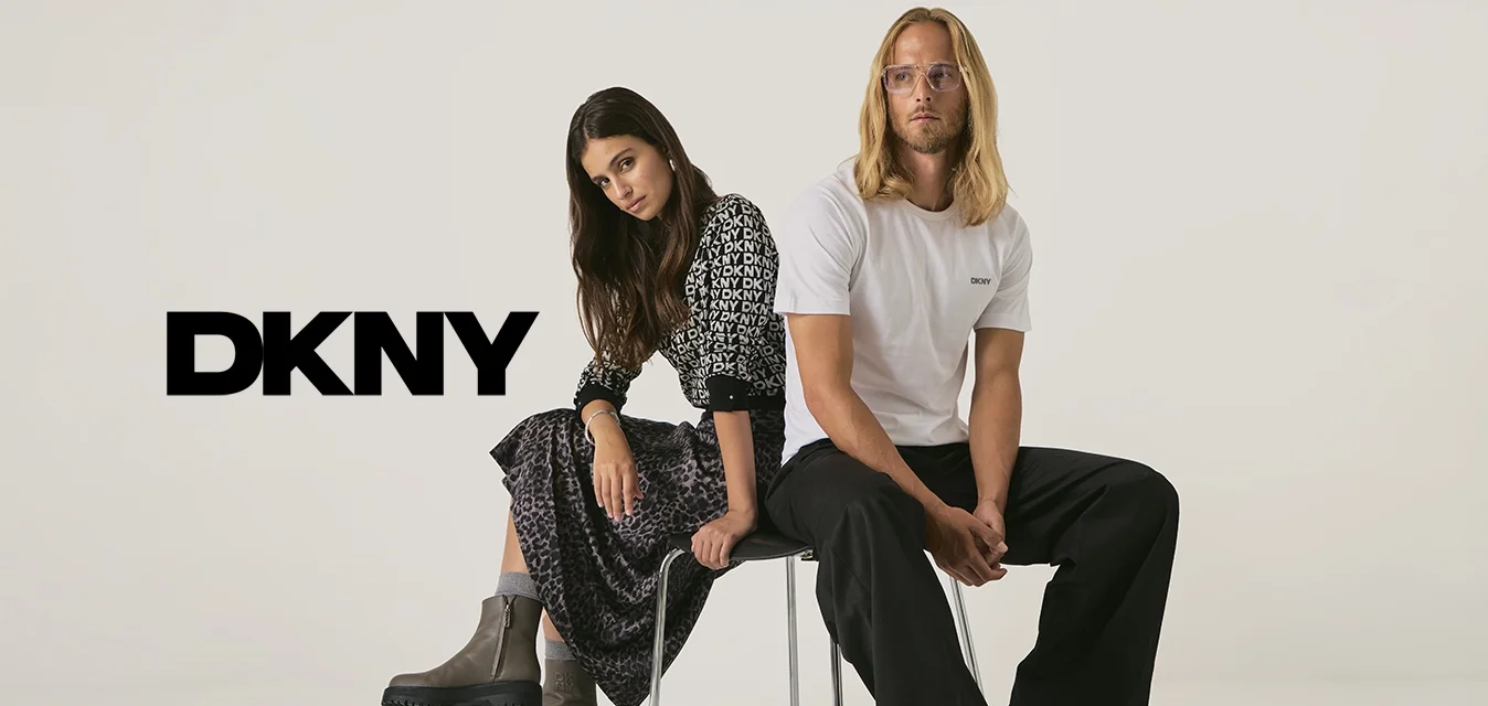 Una donna in un vestito a fantasia e stivali e un uomo in una maglietta bianca DKNY e pantaloni neri sono seduti schiena contro schiena su una sedia contro uno sfondo semplice.