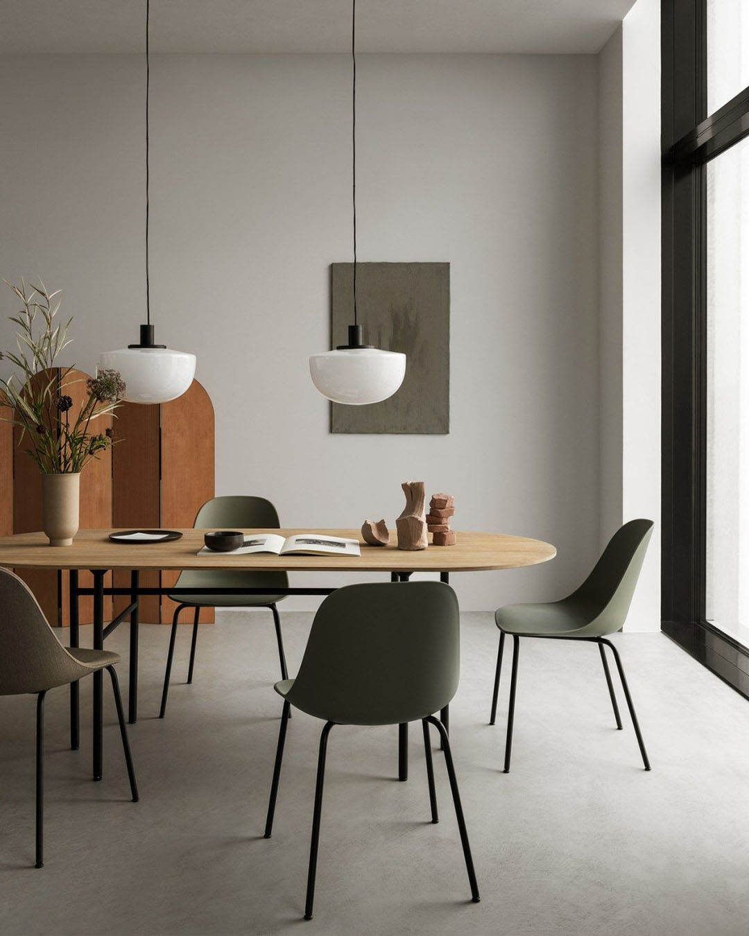 Minimalist dining room with oval wooden table, olive-green chairs, two pendant lights, decorative vases and a large floor-to-ceiling window