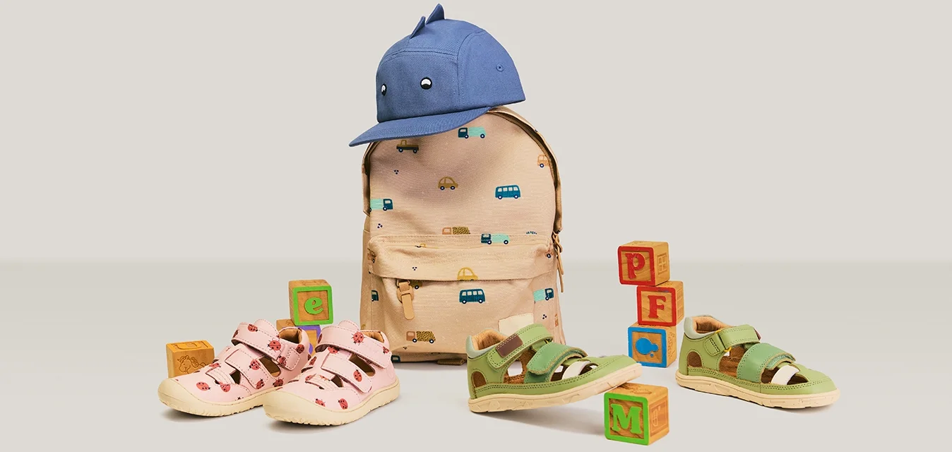 Zaino beige per bambini con stampe di veicoli, cappellino da baseball blu sopra, sandali rosa e verdi e blocchi alfabetici in legno disposti nelle vicinanze.