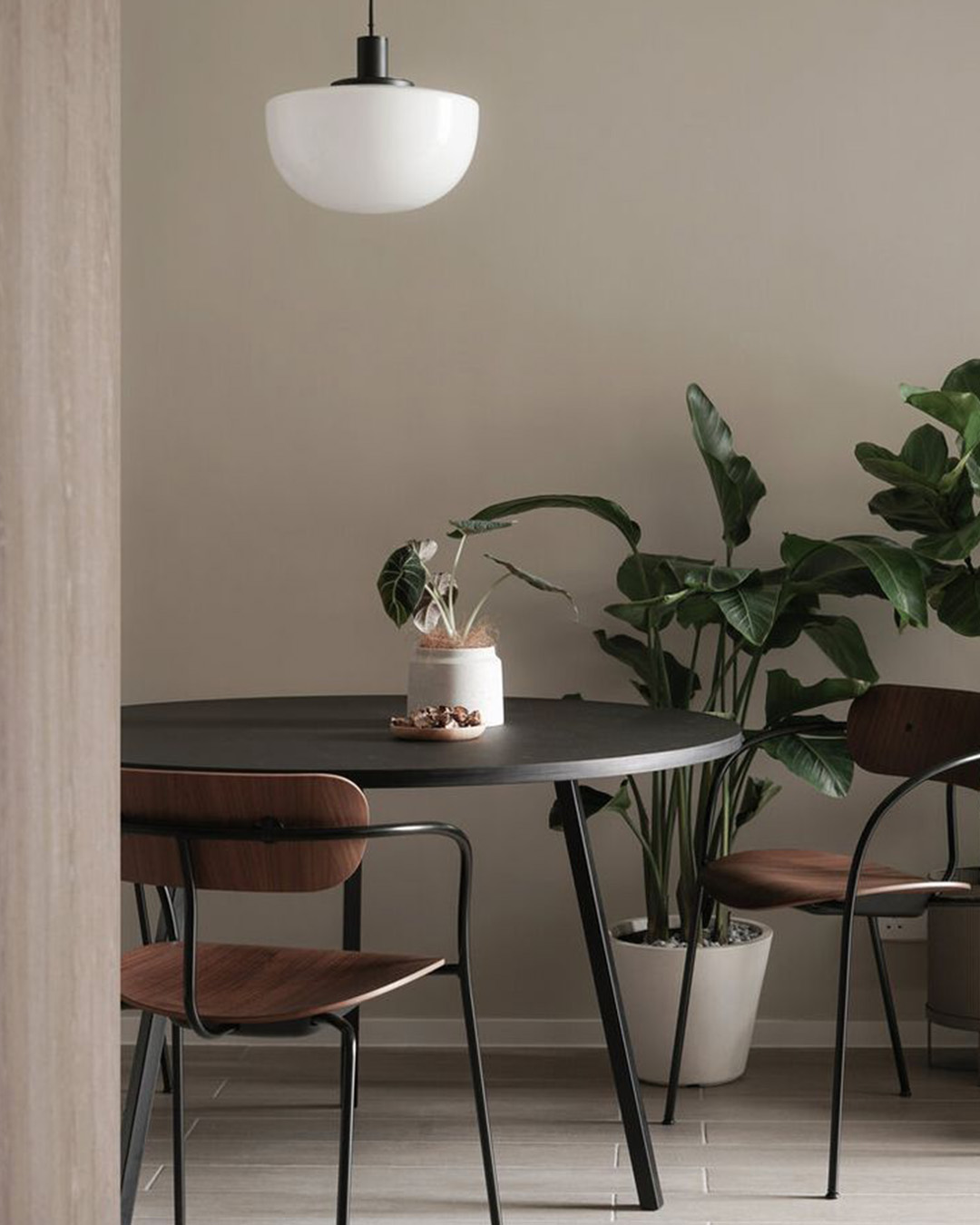 Minimalist dining area with round black table, two wooden chairs, hanging pendant light and potted indoor plants
