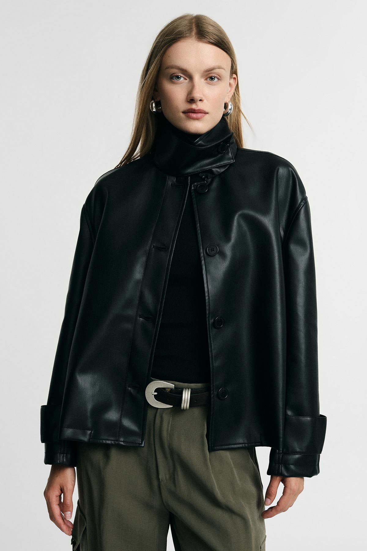 Woman wearing black leather jacket and roll-neck jumper with khaki trousers and silver belt buckle, neutral studio background