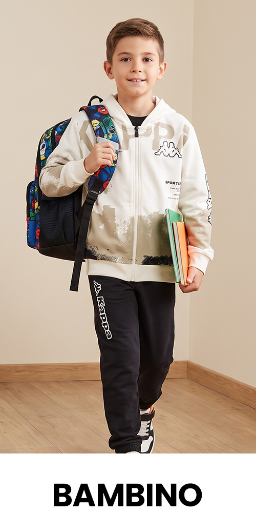 Smiling schoolboy in a white zip-up hoodie and black tracksuit, carrying a colourful backpack and books, standing indoors