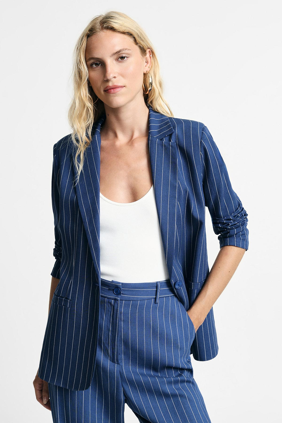 Woman wearing navy blue pinstripe blazer and matching trousers with white camisole, hand in pocket, neutral studio background