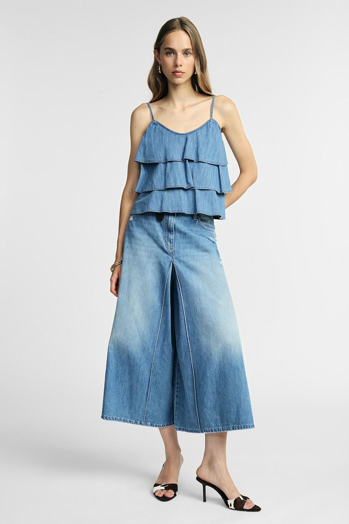 Woman modelling light-blue denim tiered cami top with wide-leg culotte jeans and black heeled mules on a plain background