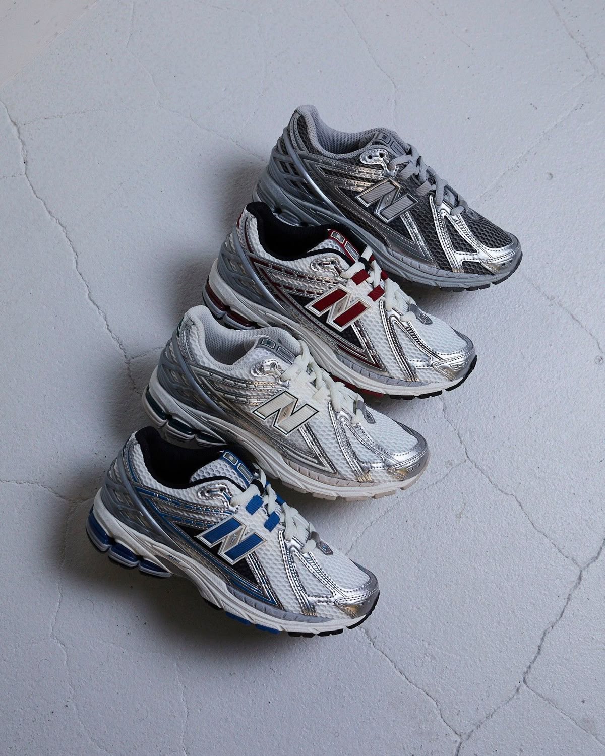 Four pairs of silver New Balance running trainers with blue, white, red and grey accents, arranged diagonally on a cracked pale concrete floor
