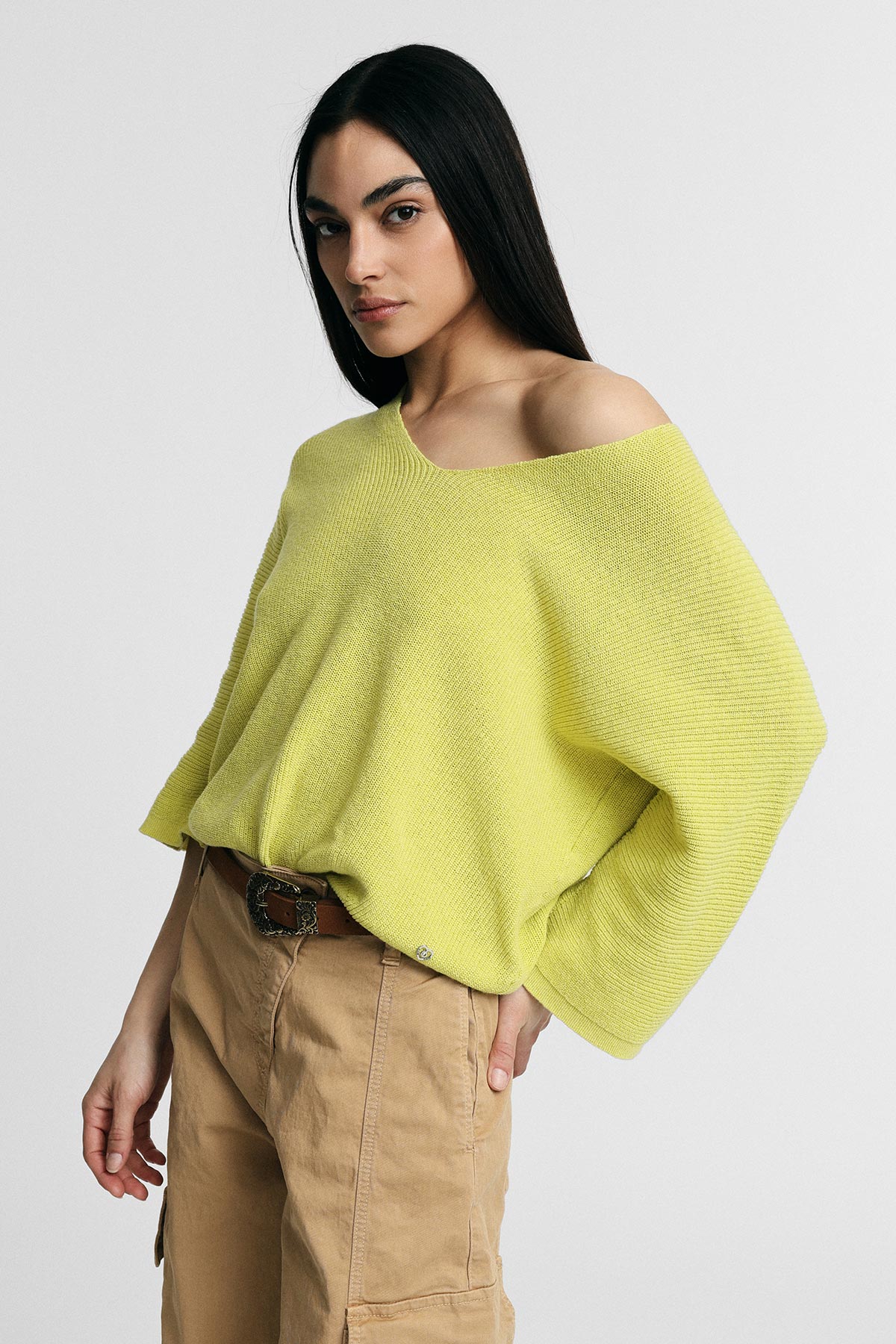 Model wearing lime-green off-the-shoulder knit sweater and beige cargo trousers with belt, posing against a plain grey background