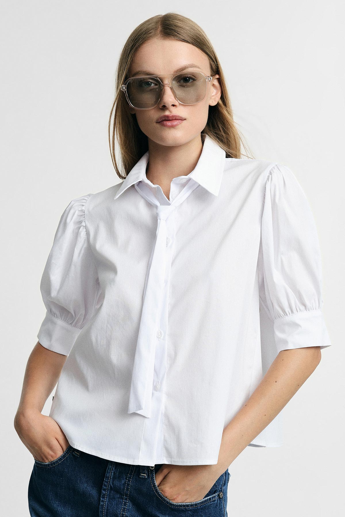 Young woman in oversized white blouse with thin necktie, sunglasses and hands in jean pockets