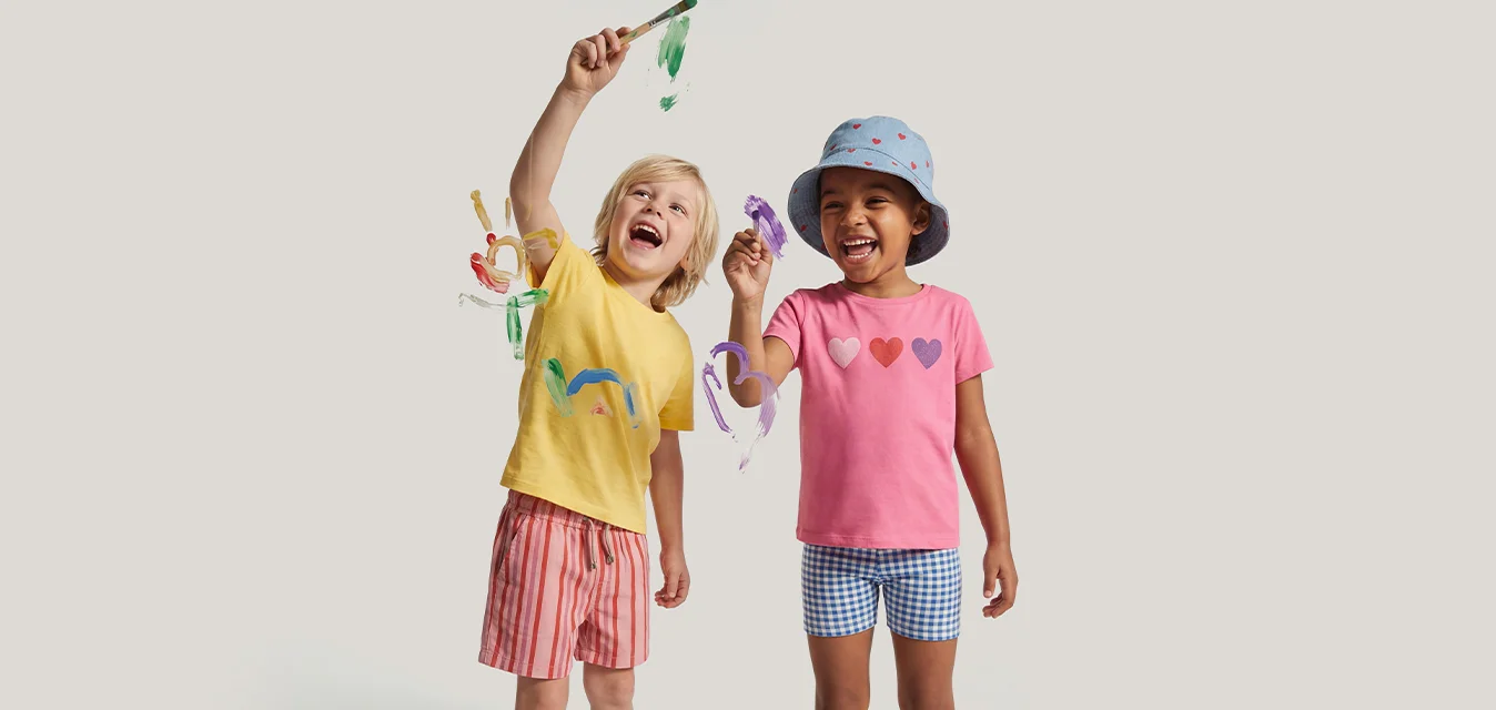 Due bambini piccoli sorridono tenendo in mano girandole colorate, uno indossa una maglietta gialla e pantaloncini rosa, l'altro una maglietta rosa, cappello blu e pantaloncini.