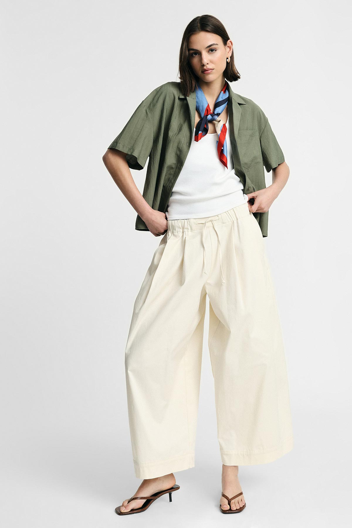 Woman modelling casual outfit: olive short-sleeve shirt over white tank, cream wide-leg trousers, silk neckerchief and heeled sandals, hands in pockets