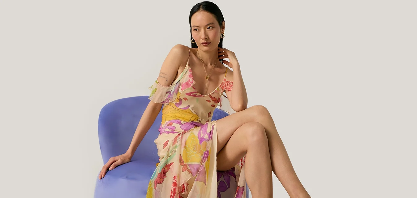 Donna che indossa un vestito floreale trasparente e sandali rossi con tacco alto, seduta su una sedia moderna blu su sfondo semplice.