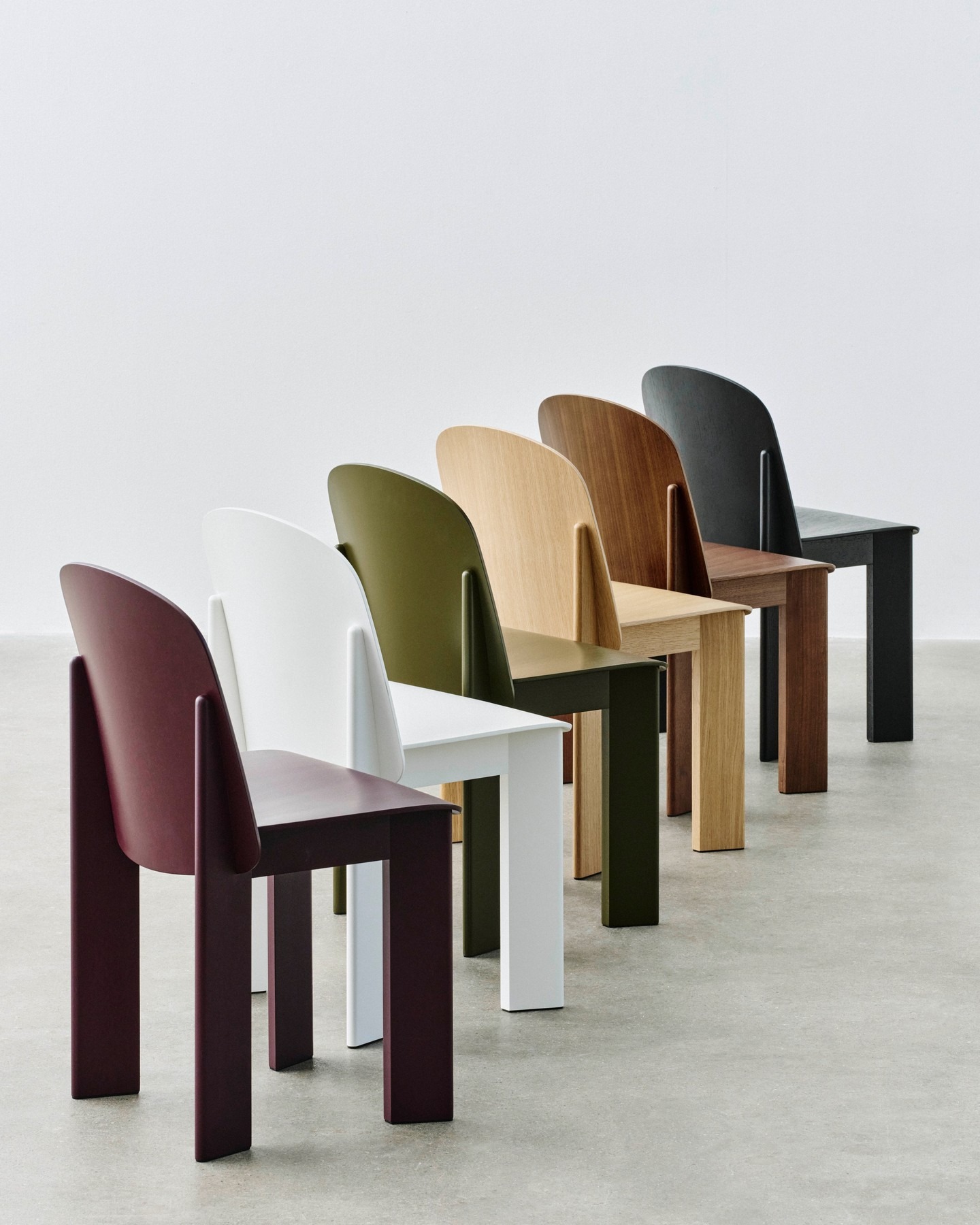 Six modern wooden dining chairs in burgundy, white, olive, light oak, walnut and black arranged in a diagonal row on a concrete floor against a white wall