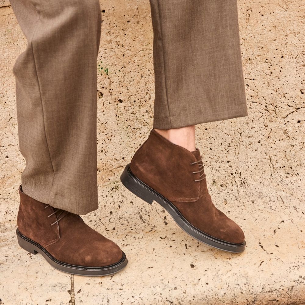Brown suede chukka boots with black sole worn with brown dress trousers on textured stone surface