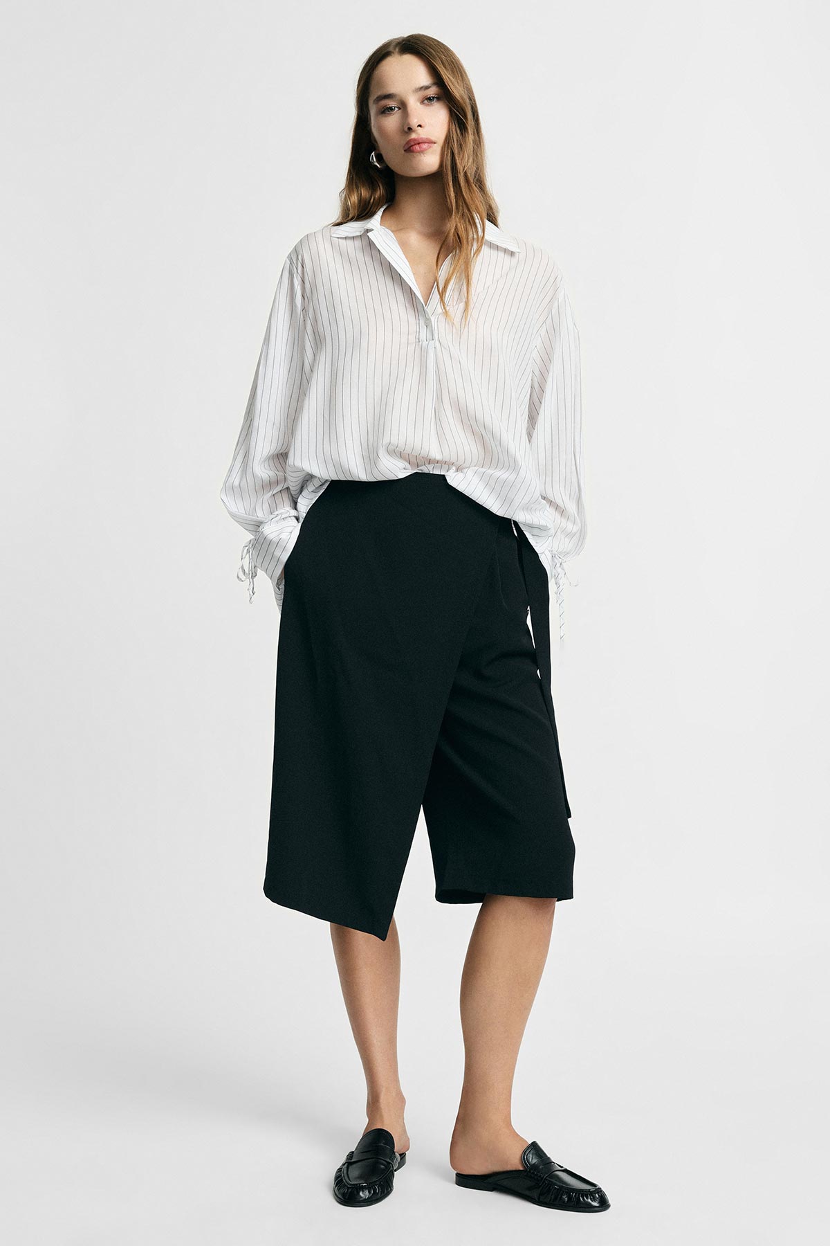 Woman modelling white pinstripe blouse tucked into black asymmetric knee-length culottes, wearing black loafers on a plain background