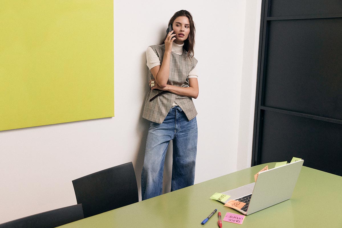 Woman on phone in a modern office, leaning against a wall in a checked waistcoat and jeans beside a laptop with sticky notes
