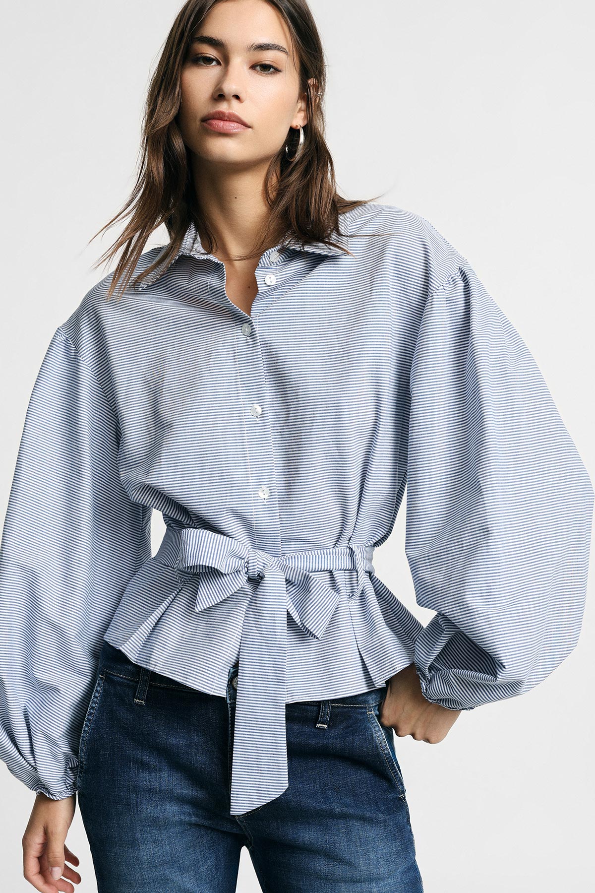 Female model wearing a blue-and-white striped belted blouse with voluminous balloon sleeves and dark jeans on a light background