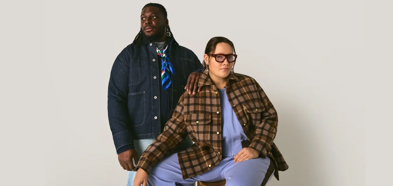 Uomo in giacca di denim scuro con foulard accanto a una donna in giacca a quadri e outfit di un leggero viola seduta su uno sgabello contro uno sfondo neutro.